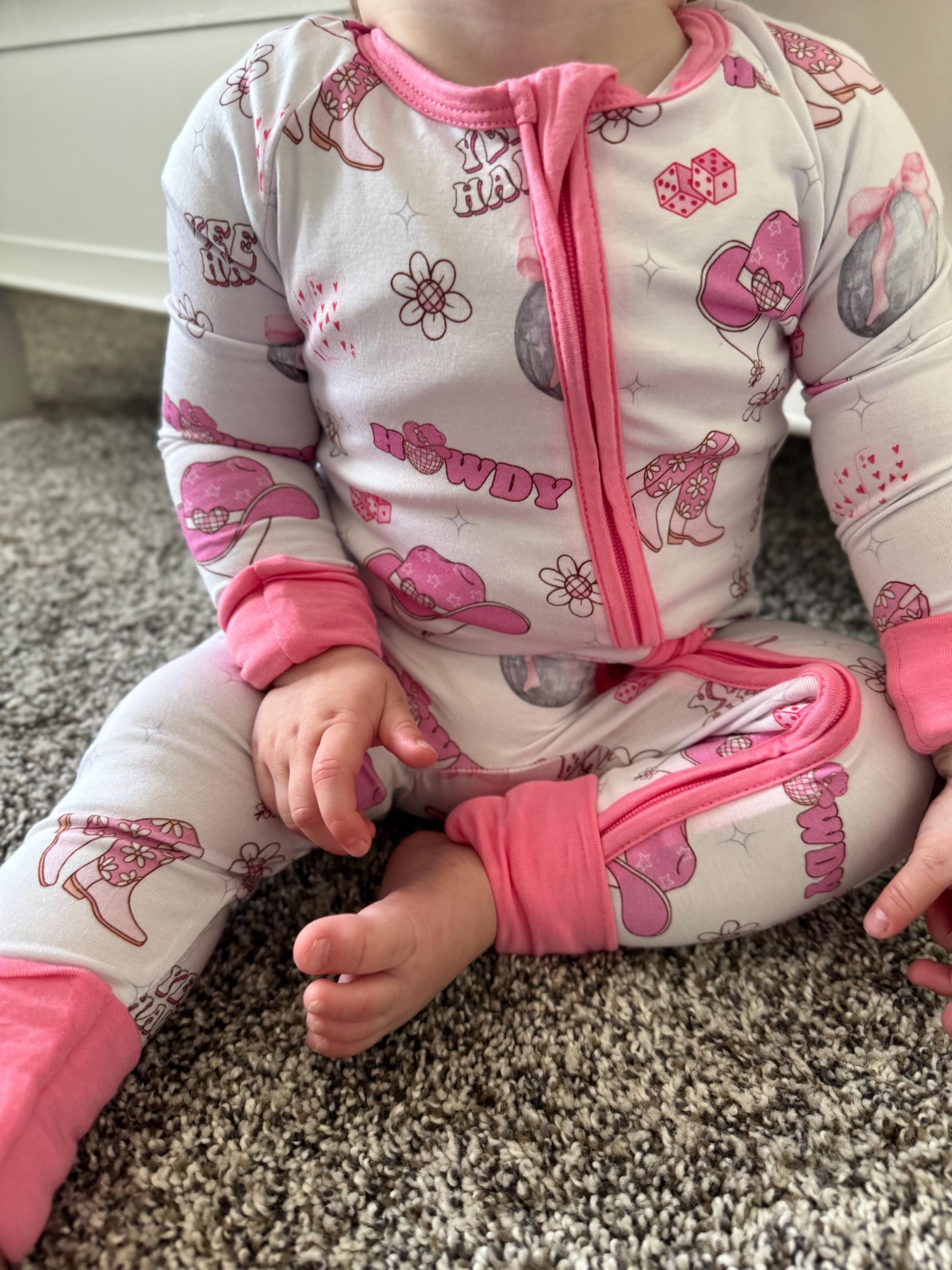 Baby in pink and white pajamas with 'Howdy' text and graphics on a carpeted floor. Bamboo convertible baby pajama with Pink and white baby pajamas with floral pattern, cowgirl pattern, western pattern, dice pattern, disco pattern, yeehaw pattern, Nashville pattern, Tennessee pattern, card pattern. 