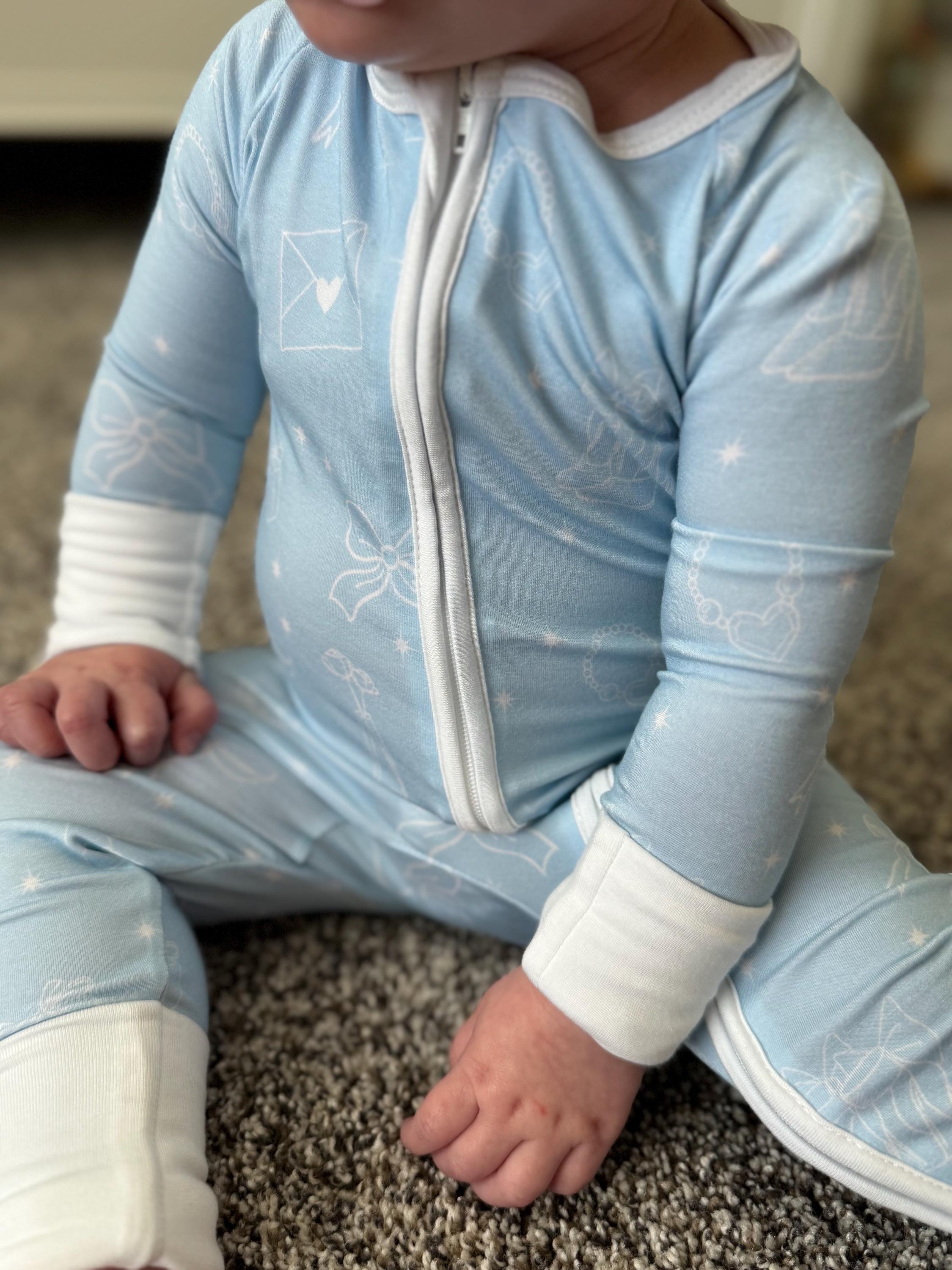 Child wearing bamboo baby clothes with light blue background and white bows, hearts, flowers and shoes. Convertible foot and mittens to allow child to grow. 
