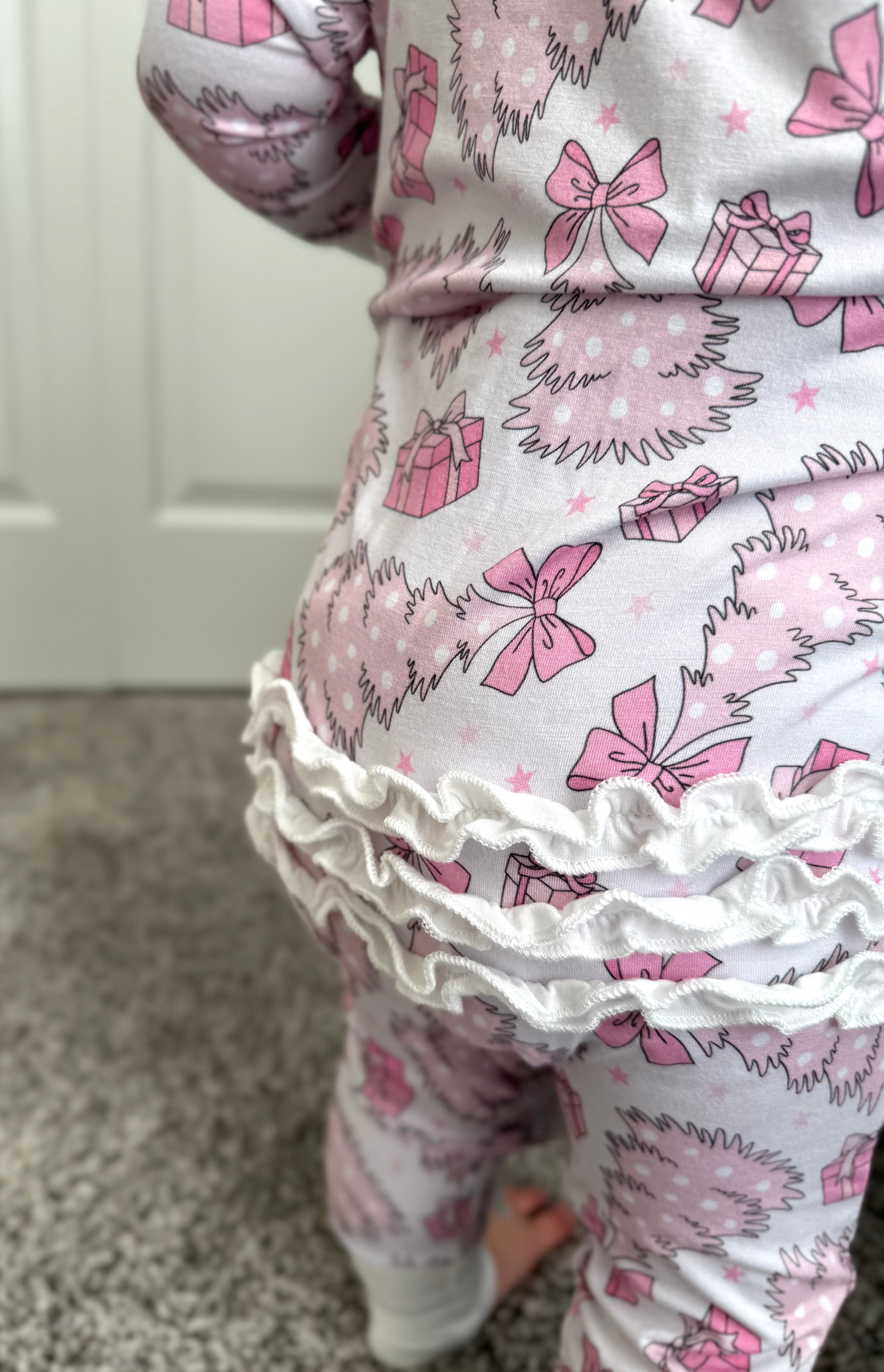 Child wearing christmas pajamas with ruffle bottom and ruffle zipper. Baby Bamboo convertible footie pajama that is pale pink with pink and white christmas trees, bows, presents and stars. Pajamas are super soft, great for sensitive skin and will grow with your little - the mittens and feet are adjustable!