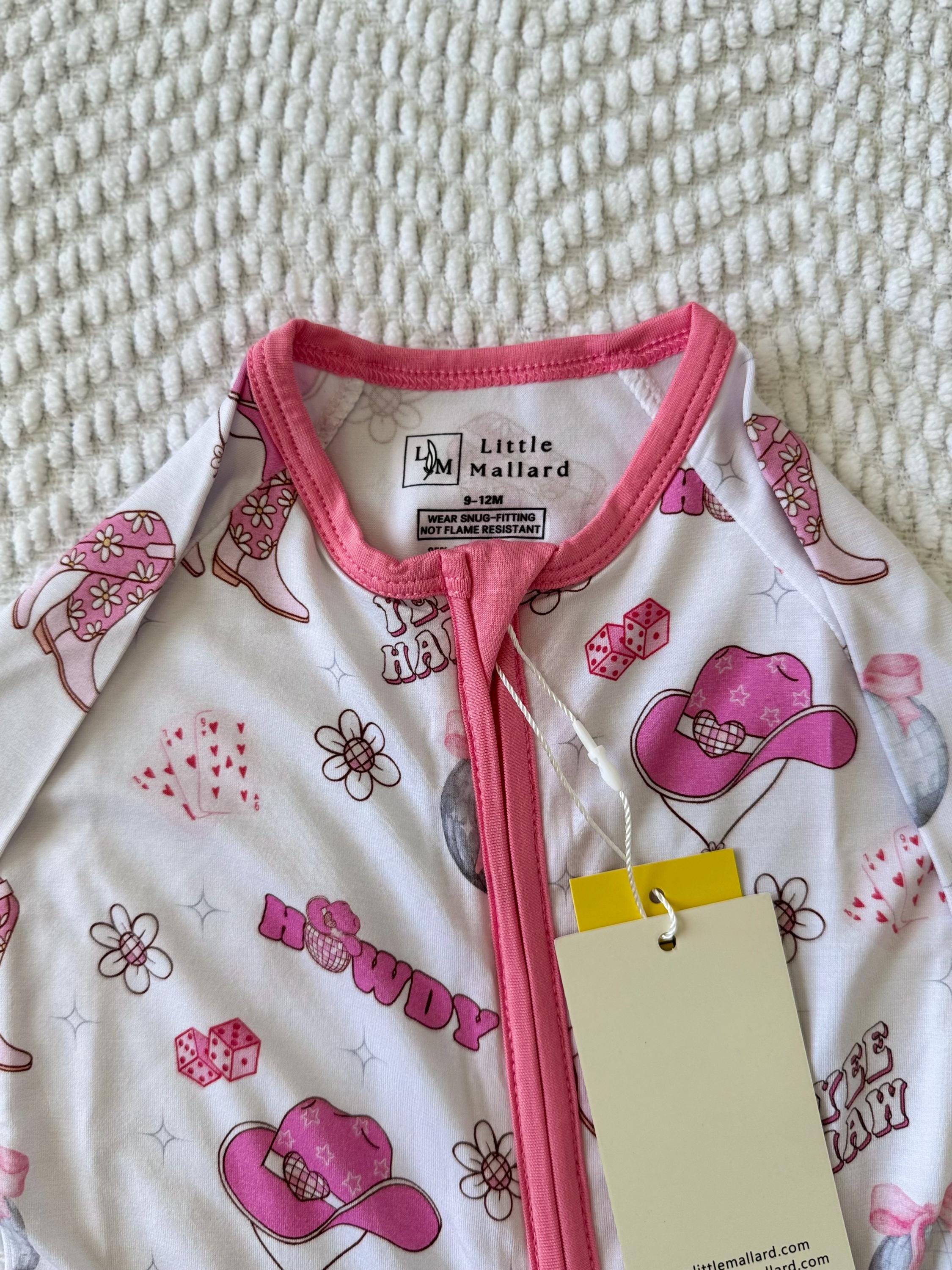 Baby onesie with pink cowboy hats and 'Howdy' text on a textured surface, brand 'Little Mallard' visible. Bamboo convertible baby pajama with Pink and white baby pajamas with floral pattern, cowgirl pattern, western pattern, dice pattern, disco pattern, yeehaw pattern, Nashville pattern, Tennessee pattern, card pattern. 